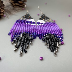 May include: Handmade beaded earrings with a striking colour gradient. The earrings feature a fringe design with a blend of black, silver, purple, and white beads. A unique accessory for any outfit.