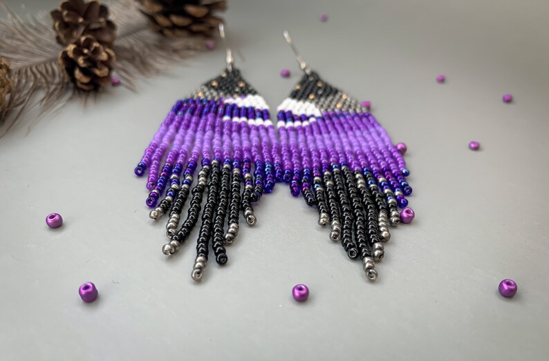 May include: Handmade beaded earrings with a striking colour gradient. The earrings feature a fringe design with a blend of black, silver, purple, and white beads. A unique accessory for any outfit.
