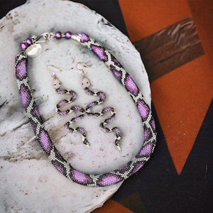 Pode incluir: Um colar de cobra de contas rosa e branco com brincos de cobra combinando. O colar e os brincos são feitos de contas pequenas e têm uma aparência texturizada, semelhante à de uma cobra.