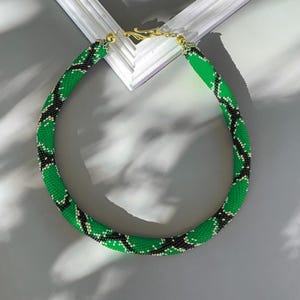 Puede incluir: Un collar de cuentas verdes y negras. El collar tiene un diseño en espiral y está hecho de pequeñas cuentas. El cierre es de color dorado. El collar se muestra sobre una superficie gris.