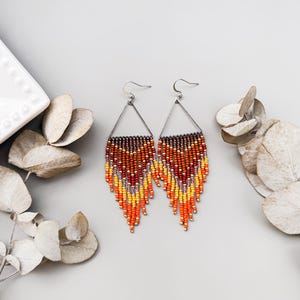 Pode incluir: Um par de brincos de contas com uma armação triangular prateada. Os brincos apresentam um gradiente de cores, incluindo castanho, laranja, amarelo e vermelho. A franja de contas pende, criando um acessório elegante e colorido.