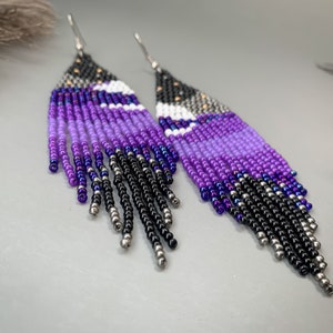 May include: A pair of handmade beaded earrings. The design incorporates a gradient of colours, including black, silver, and shades of purple. The top section is black with silver beads, fading into purple and white, and ending with black fringe.