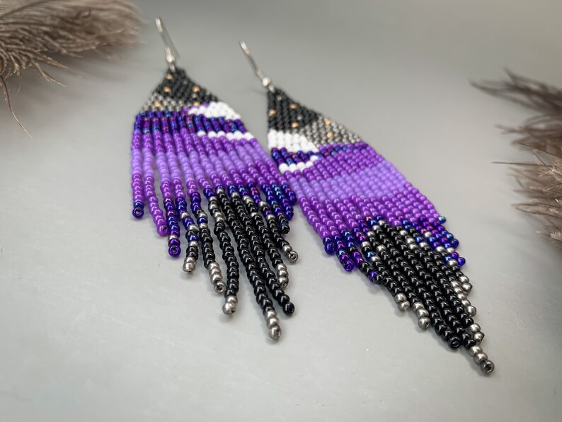 May include: A pair of handmade beaded earrings. The design incorporates a gradient of colours, including black, silver, and shades of purple. The top section is black with silver beads, fading into purple and white, and ending with black fringe.