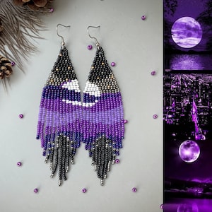 May include: Handmade beaded earrings in shades of purple, black, silver, and white. The earrings feature a geometric design with a fringe at the bottom. The earrings are designed with a hook closure. The earrings are a unique accessory.
