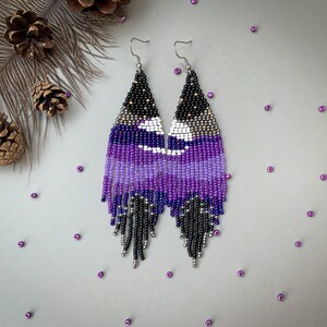 May include: Handmade beaded earrings in a triangular shape, featuring a night sky design. The earrings transition from black at the top to shades of purple and white, with silver fringe. The earrings are approximately 10 cm long.