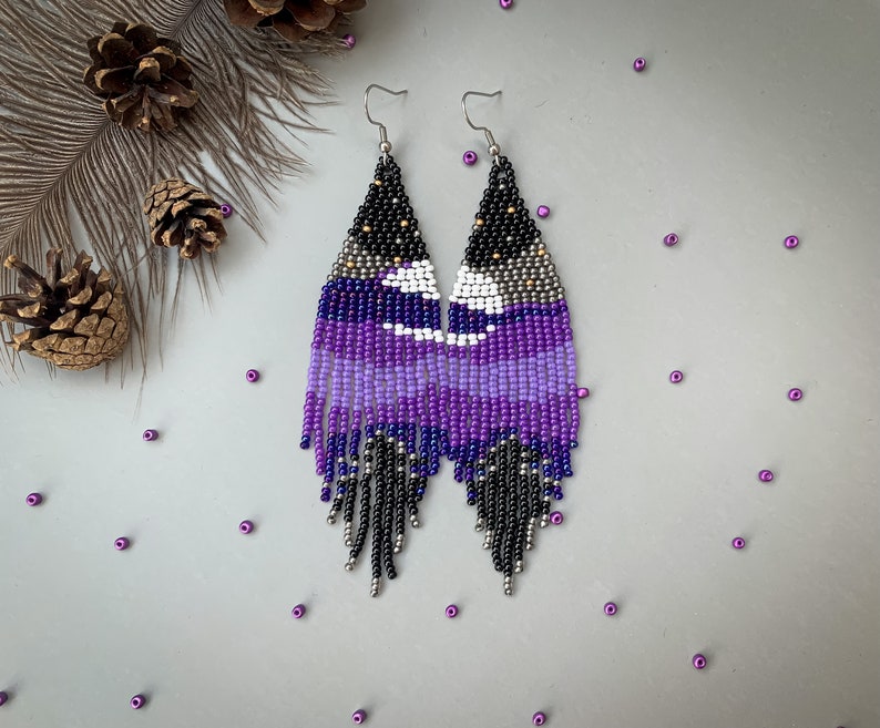 May include: Handmade beaded earrings in a triangular shape, featuring a night sky design. The earrings transition from black at the top to shades of purple and white, with silver fringe. The earrings are approximately 10 cm long.