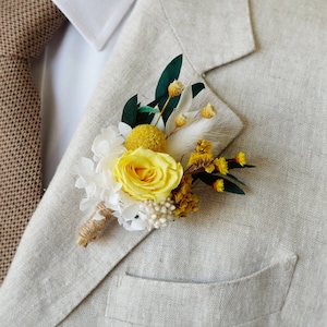 May include: A yellow rose, white hydrangea, and yellow billy balls are arranged in a boutonniere with greenery and white feathers.