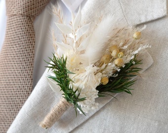 Puede incluir: Un boutonniere de flores secas blancas y beige con follaje verde, atado con cuerda.