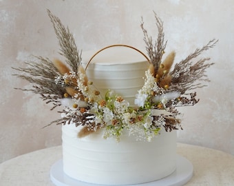 May include: A two-tiered white wedding cake decorated with dried flowers and pampas grass. A gold metal hoop encircles the top tier of the cake.