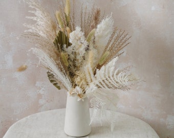 Può includere: Un'arrangiamento di fiori secchi in un vaso di ceramica bianco. Il bouquet presenta una varietà di erbe, felci e altre piante secche di colore beige, crema e marrone chiaro. Il vaso è cilindrico con una parte superiore leggermente svasata.