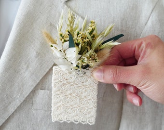 Puede incluir: Una chaqueta beige claro con tejido texturizado, combinada con una corbata estampada. Un pequeño arreglo floral con flores secas y vegetación está sujeto a un pañuelo de bolsillo de encaje crema.