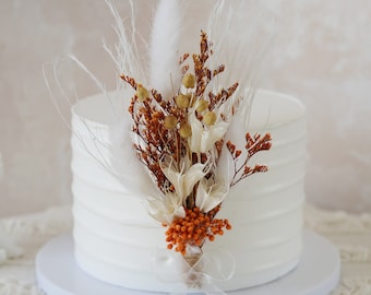 May include: A white cake with a decorative topper of dried flowers and grasses in shades of brown, white, and orange.