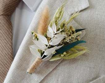 Puede incluir: Un boutonniere de flores secas blancas y verdes con un lazo de cuerda marrón. El boutonniere está prendido a una chaqueta de lino marrón claro.