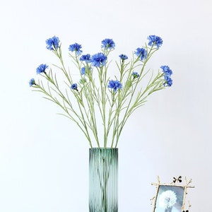 May include: A bouquet of blue artificial flowers in a green glass vase with vertical lines. The vase is on a dark surface with a gold picture frame.