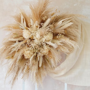 May include: A large dried floral arrangement with pampas grass, dried flowers, and wheat in beige and cream tones. The bouquet is artfully arranged and displayed with cream fabric draped over a white arched structure.