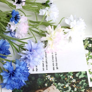 May include: A bouquet of artificial flowers with blue, white, and pink blossoms. The flowers are arranged in a vase and are surrounded by green leaves.