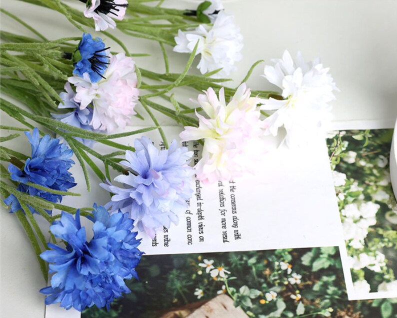 May include: A bouquet of artificial flowers with blue, white, and pink blossoms. The flowers are arranged in a vase and are surrounded by green leaves.