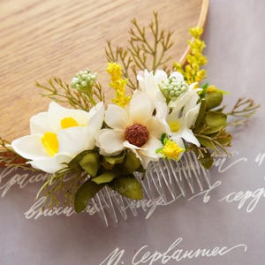 May include: A floral hair comb featuring white and yellow flowers with green leaves and accents. The comb has a clear plastic base and is adorned with small yellow and green buds. The flowers are arranged on a wooden embroidery hoop.