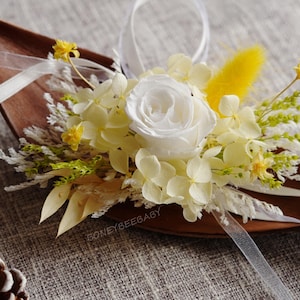 Peut inclure: Composition florale avec une rose blanche, des touches de jaune et des rubans blancs, présentée sur une base brune en forme de feuille. L'arrangement comprend des fleurs et de la verdure blanches et jaunes, créant une esthétique délicate et naturelle.