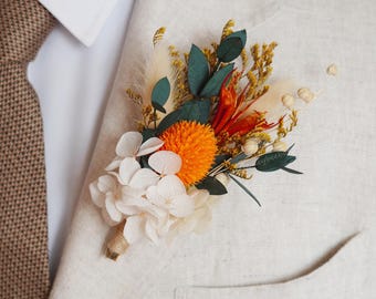 Puede incluir: Un prendido floral con una vibrante flor naranja, flores blancas y acentos verdes y amarillos. El arreglo está sujeto a una chaqueta de traje de color claro, combinada con una corbata marrón estampada. Un accesorio elegante para ocasiones especiales.