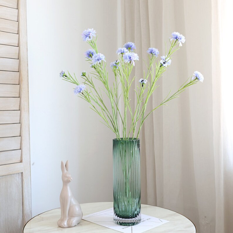 May include: A bouquet of light blue artificial flowers in a green glass vase with a ribbed design. The vase is on a white table with a small ceramic rabbit figurine.