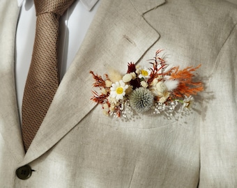 May include: A beige linen suit jacket with a brown patterned tie and a small bouquet of dried flowers pinned to the lapel. The bouquet includes white daisies, brown and orange dried grasses, and a small gray thistle.