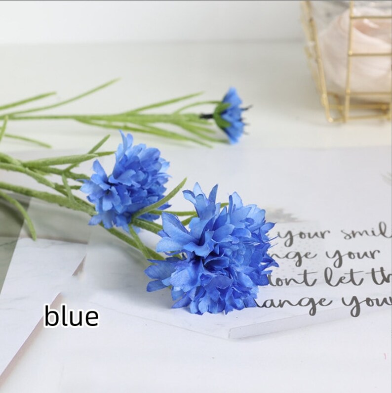 May include: A bouquet of blue artificial flowers with green stems. The flowers are arranged in a cluster and have a delicate, ruffled appearance.