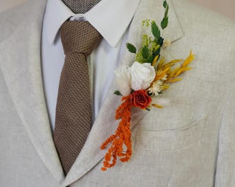 Puede incluir: Una blazer beige claro con una corbata marrón texturizada y una camisa blanca. Un prendido floral con una rosa blanca, acentos naranjas y amarillos está prendido a la solapa de la blazer. El prendido añade un toque de color.