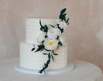 May include: A two-tiered white wedding cake with buttercream frosting and a simple, elegant design. The cake is decorated with a cascading arrangement of white flowers and green leaves.