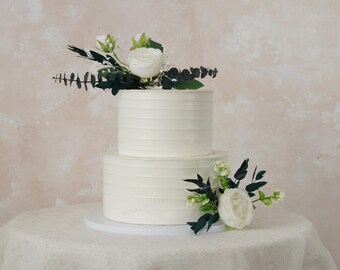 May include: A two-tiered white wedding cake decorated with simple lines and topped with white flowers and green leaves.