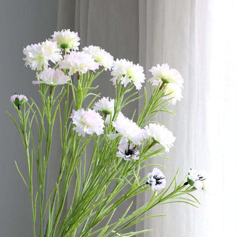 May include: A bouquet of artificial white flowers with green stems. The flowers have a delicate, ruffled appearance and are arranged in a vase.