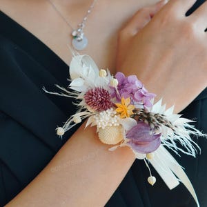 May include: A wrist corsage featuring a cluster of dried flowers in shades of purple, white, yellow, and burgundy. The floral arrangement includes feathers and is secured with white ribbons. The corsage is worn on a wrist.