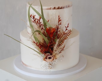 May include: A two-tiered white frosted cake decorated with dried flowers and greenery. The cake is adorned with orange, brown, and green foliage, including pampas grass and a small rose. The cake sits on a white cake stand.