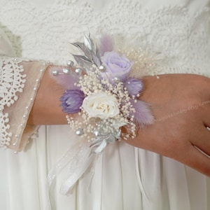 May include: A wrist corsage featuring a white rose, lavender flowers, and silver accents. The floral arrangement includes small white buds, silver leaves, and delicate ribbons. The corsage is designed for a special occasion, such as a wedding or prom.