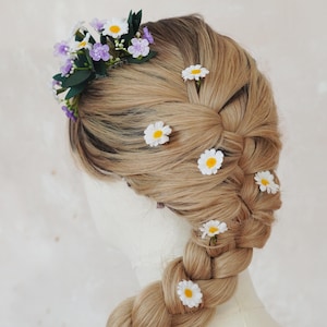 May include: A blonde braid adorned with small white daisies and a floral crown of purple flowers and green leaves. The hairstyle is displayed on a mannequin head, showcasing a romantic and natural aesthetic, perfect for weddings or special events.