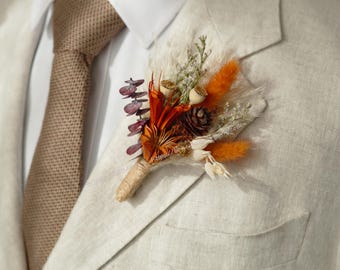 May include: A tan suit jacket with a brown patterned tie and a small bouquet of dried flowers pinned to the lapel. The bouquet includes orange, white, and brown flowers.