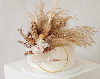 May include: A two-tiered white cake decorated with dried pampas grass, wheat, and delicate white and pink flowers. A gold hoop accents the front. The cake sits on a white pedestal.