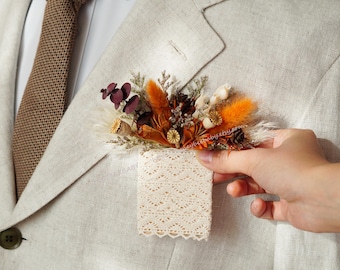 May include: A small bouquet of dried flowers in shades of orange, brown, and white, arranged in a delicate lace pocket. The bouquet is being held by a hand, ready to be pinned to a garment.