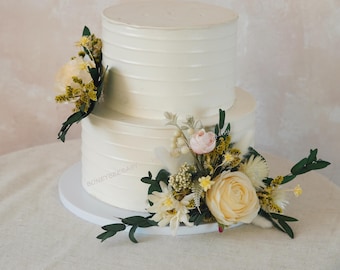 Op de afbeelding: Een witte bruidstaart met twee lagen, versierd met botercrème en een bloemstuk met crème, gele en roze bloemen.