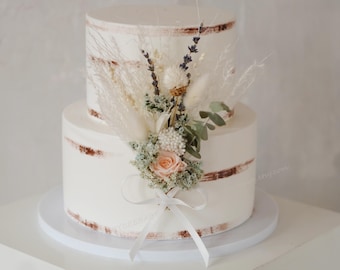 May include: A two-tiered white cake with a rustic design, adorned with dried flowers and a delicate ribbon. The cake features a semi-naked style with visible brown frosting accents. The floral arrangement includes a peach rose and pampas grass.