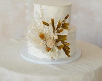 May include: A two-tiered white wedding cake decorated with dried flowers. The flowers are a mix of white and beige pampas grass, dried wheat, and small white and beige flowers.