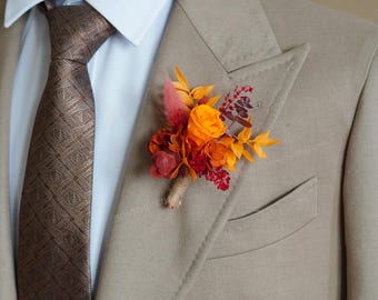 Puede incluir: Un blazer color beige con una corbata marrón estampada y un prendido floral. El prendido presenta flores y follaje de color naranja, rojo y amarillo. El blazer tiene una camisa azul claro debajo.