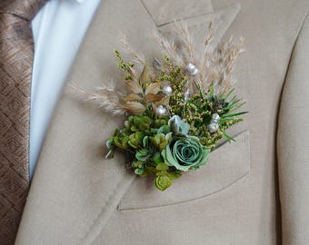May include: A boutonniere featuring a green rose, succulents, and dried grasses, accented with silver beads. The floral arrangement is pinned to a tan suit jacket, paired with a patterned brown tie.