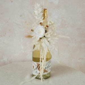 Puede incluir: Una botella de champán decorada con flores secas blancas y vegetación. La botella está envuelta en tul blanco y tiene una cinta dorada atada alrededor del cuello.