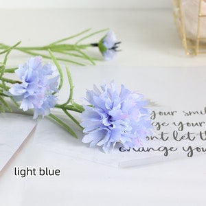 May include: Light blue artificial flower stems with multiple flower heads. The flowers are made of fabric and have a delicate, ruffled appearance.
