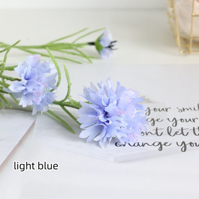May include: Light blue artificial flower stems with multiple flower heads. The flowers are made of fabric and have a delicate, ruffled appearance.