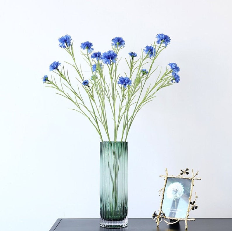 May include: A bouquet of blue artificial flowers in a green glass vase with vertical lines. The vase is on a dark surface with a gold picture frame.