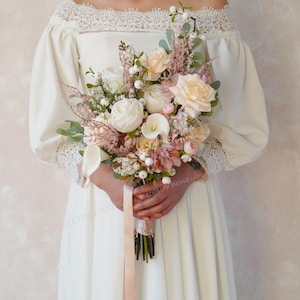 May include: A bridal bouquet featuring a mix of white, cream, and blush-colored flowers. The arrangement includes roses, calla lilies, and other delicate blooms, tied with a blush ribbon. The bouquet is held against a white dress.