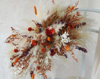 May include: A dried floral arrangement featuring fluffy, cream-colored pampas grass, accented with orange, red, and white dried flowers and foliage. The arrangement is artfully composed, creating a visually appealing display. The overall theme is rustic and natural.