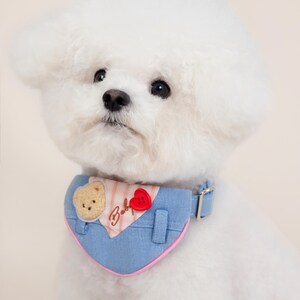 May include: A white Bichon Frise dog wearing a blue and pink bandana with a bear and heart applique. The bandana has the word "Baby" embroidered on it.